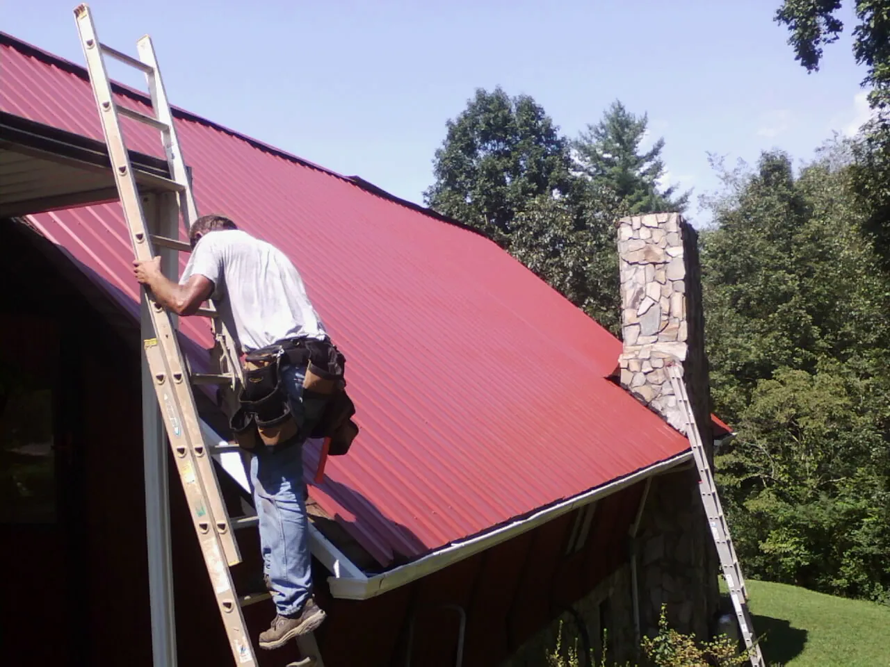 Roofer on a ladder beside a red metal roof and stone chimney — Soffit & Fascia Repair by Roofing Brothers in Clarendon